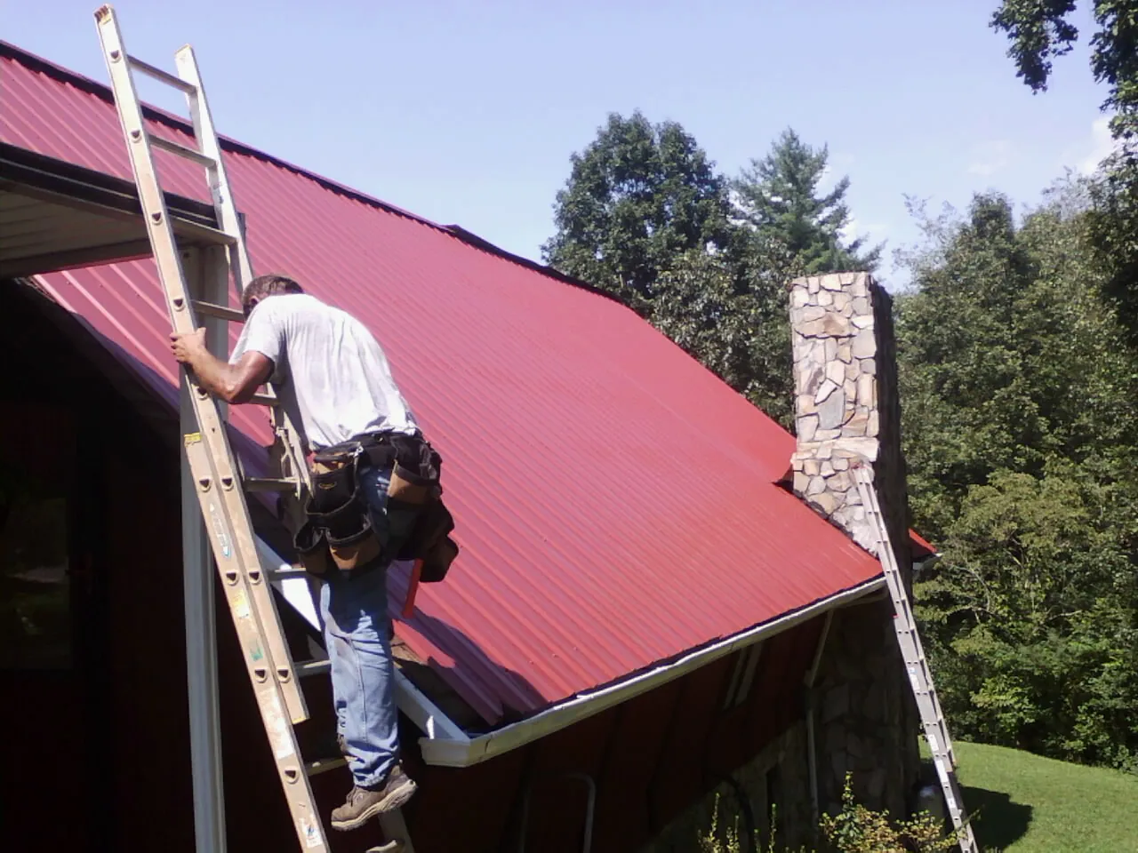 Roofer on a ladder beside a red metal roof and stone chimney — Soffit & Fascia Repair by Roofing Brothers in Mount Rainier